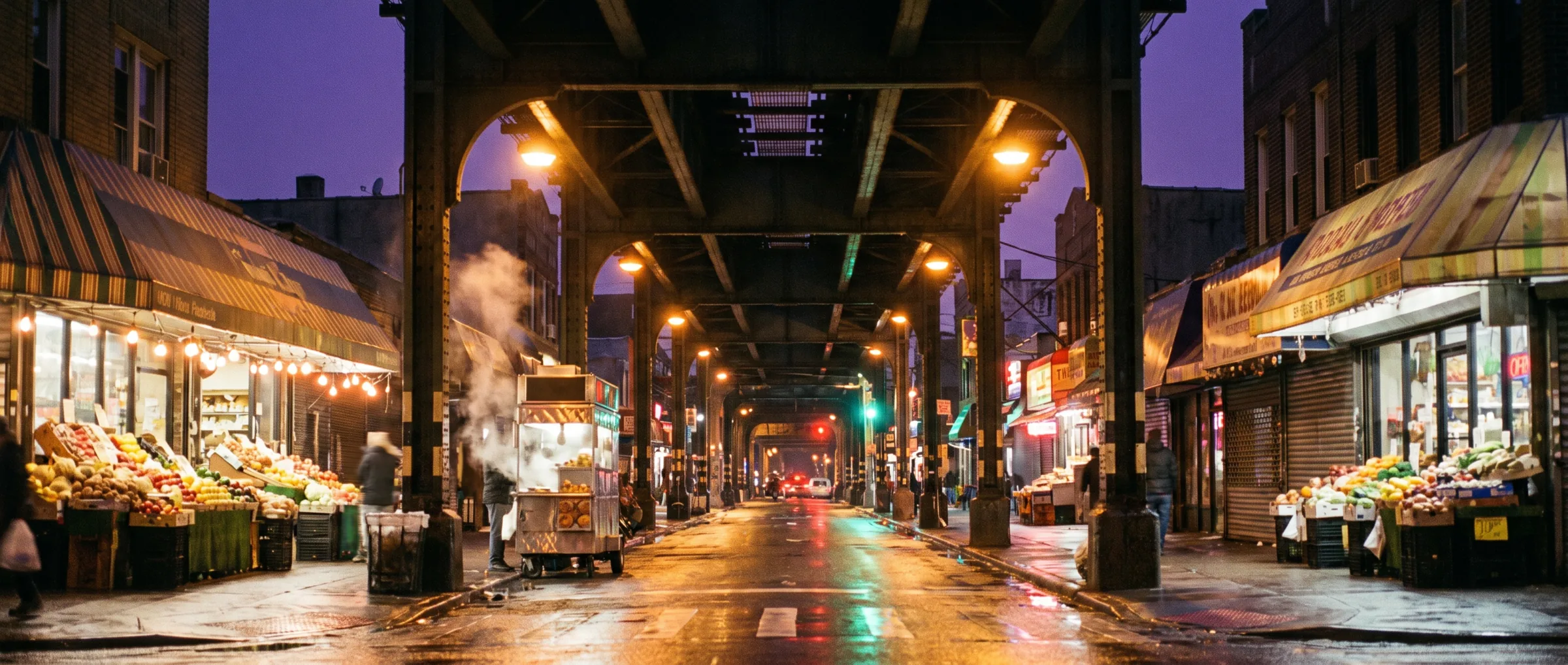 Street at night under an elevated train line, awnings and market stalls lit by sodium lamps